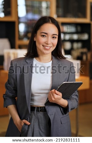 Young smiling successful professional leader Asian business woman, female executive manager, saleswoman wearing suit holding digital tablet standing in office looking at camera, vertical portrait.