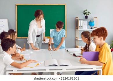 Young smiling teacher is engaged with student standing at table against the background of blackboard, other children are laughing while sitting at the table and making notes in their school notebooks. - Powered by Shutterstock