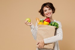 Young Smiling Vegetarian Cheerful Woman 20s In Casual Clothes Hold Paper Bag With Vegetables Holding Apple Fruit Eating Isolated On Plain Pastel Beige Background Studio Portrait. Shopping Concept.