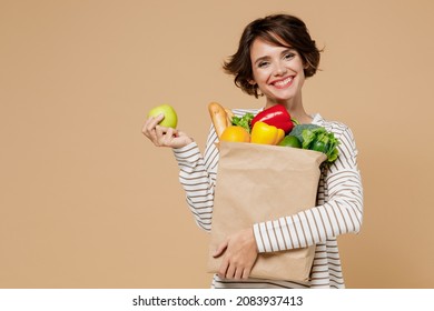 Young smiling vegetarian cheerful woman 20s in casual clothes hold paper bag with vegetables holding apple fruit eating isolated on plain pastel beige background studio portrait. Shopping concept. - Powered by Shutterstock