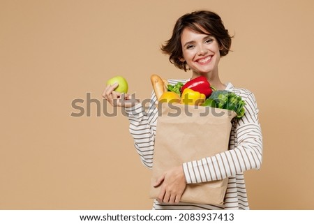 Young smiling vegetarian cheerful woman 20s in casual clothes hold paper bag with vegetables holding apple fruit eating isolated on plain pastel beige background studio portrait. Shopping concept.