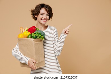 Young smiling vegetarian woman 20s in casual clothes hold paper bag with vegetables after shopping in greengrocery point finger aside on workspace area isolated on plain pastel beige background studio