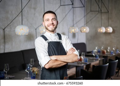 A young, smiling waiter in a restaurant, standing next to the tables with a glass of wine. He wears an apron, looks confidently, folded his arms over his chest