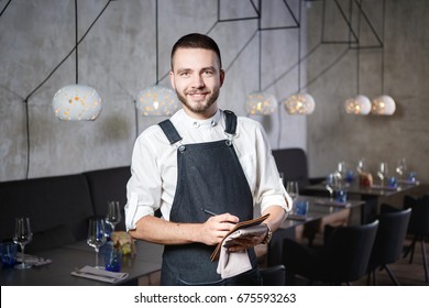 A Young, Smiling Waiter In A Restaurant, Standing Next To The Tables With A Glass Of Wine. Dressed In An Apron, Will Take An Order Holding A Notebook And A Pen