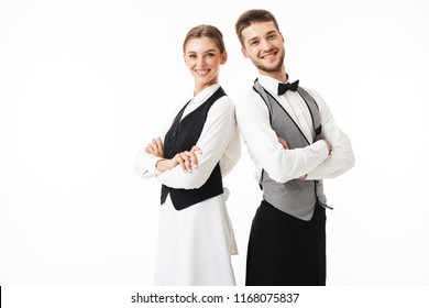 Young smiling waiter and waitress in white shirts and vests sstanding back to back joyfully looking in camera with arms folded over white background