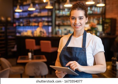 Young smiling waitress in apron and t-shirt standing in front of camera while using touchpad and meeting guests in cafe - Powered by Shutterstock