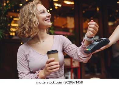 Young smiling woman 20s in casual clothes at cafe buy breakfast sit at table hold wireless bank payment terminal smart watch to process acquire payments relax in restaurant during free time indoors.