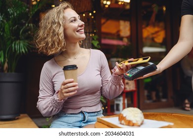 Young smiling woman 20s in casual clothes at cafe buy breakfast sit at table hold wireless bank payment terminal mobile phone to process acquire payments relax in restaurant during free time indoors.