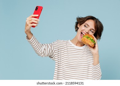 Young smiling woman 20s in casual sweater look camera hold burger doing selfie shot on mobile cell phone isolated on plain pastel light blue background studio portrait. People lifestyle food concept