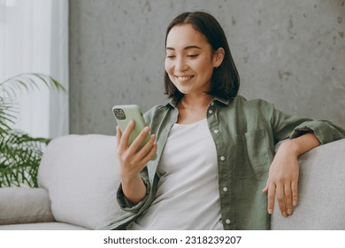 Young smiling woman of Asian ethnicity wears casual clothes hold in hand use mobile cell phone sits on grey sofa couch stay at home hotel flat rest relax spend free spare time in living room indoor