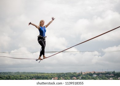 Young smiling woman balancing with arms raised on the slackline rope on the background of clear sky