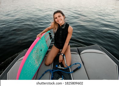 Young Smiling Woman In Black Swimsuit Sits On Boat And Holds Surf Style Wakeboard With Her Hand