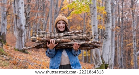 Young smiling woman with brown hat carring firewood in her hands in the forest. Preparing firewood for winter concept, collecting firewood for camping in the mountain.
