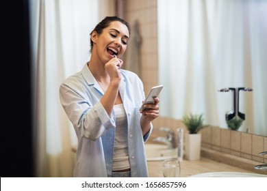 Young smiling woman brushing teeth and reading text message on mobile phone in the bathroom.  - Powered by Shutterstock