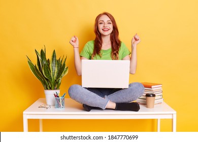 Young smiling woman in casual clothes clenching fists like winner while sitting on desk with laptop on knees, posing isolated on yellow wall, ginger happy girl near flower pot and stack of books.