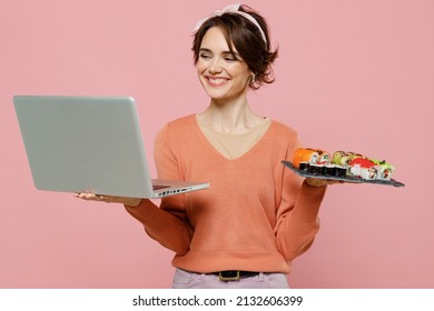 Young Smiling Woman In Casual Clothes Hold In Hand Makizushi Sushi Roll Served On Black Plate Traditional Japanese Food Hold Use Work On Laptop Pc Computer Isolated On Plain Pastel Pink Background.