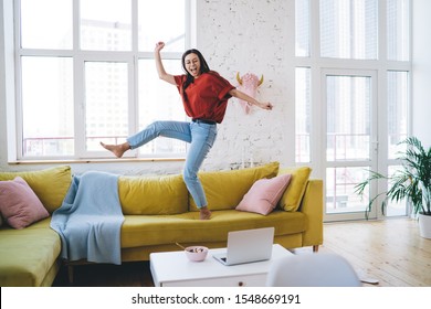 Young Smiling Woman In Casual Clothing And Glasses Dancing On Yellow Couch In Living Room Near Coffee Table With Laptop And Breakfast Bowl