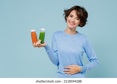 Young smiling woman in casual sweater hold pressed juice green orange vegetable smoothie as detox diet put arm on belly isolated on plain pastel light blue background. People lifestyle food concept - Shutterstock ID 2118594188