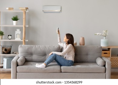 Young smiling woman in casual wear sitting on couch in living room resting holding remote controller switch on air conditioner make regulate comfort temperature at home enjoy fresh or warm air concept - Shutterstock ID 1354368299