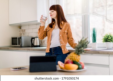 Young Smiling Woman In Casual Wear, Drinking Minearl Water, While Standing In Modern Kitchen At Home, Starting Her Active Day. Ready For Eating Healthy Breakfast And Working Online