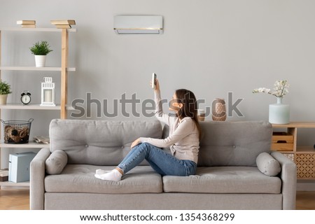 Young smiling woman in casual wear sitting on couch in living room resting holding remote controller switch on air conditioner make regulate comfort temperature at home enjoy fresh or warm air concept