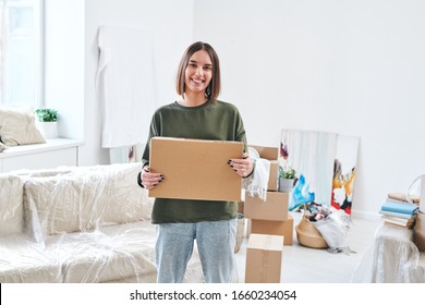 Young smiling woman in casualwear holding carton box while standing in front of camera in living-room of new flat or house