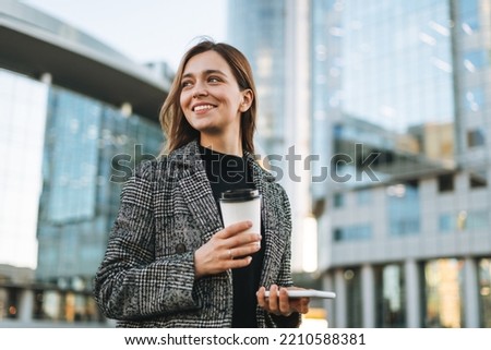 Young smiling woman in coat with coffee cup using mobile phone in evening city street