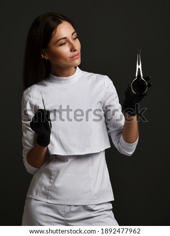 Young smiling woman doctor dentist in white uniform and latex gloves standing holding medical dental tools in hands over dark background. Dentist, stomatologist, orthopedist concept