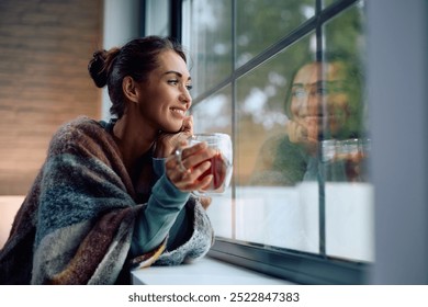 Young smiling woman drinking tea and looking through the window at home. Copy space. - Powered by Shutterstock