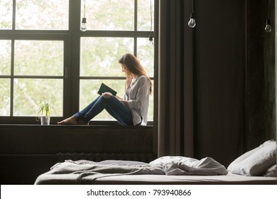 Young smiling woman enjoying new bestseller book sitting on window sill, happy book lover reading fiction literature relaxing at home, student teenager study textbook in modern cozy loft bedroom