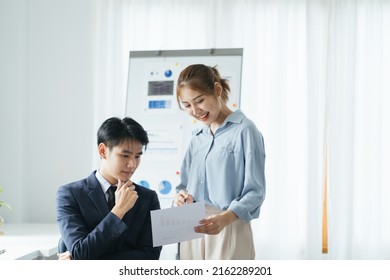 Young Smiling Woman Explaining To Serious Asian Coworker Project Strategy. Diverse Startup Coworkers Students Woman And Man Talking Discussing Working In Modern Office Using Computer.