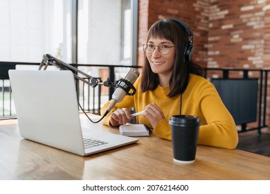 Young smiling woman in eyeglasses recording and broadcasting her podcast in studio. Female radio host in headphones recording live audio podcast using microphone and laptop, holding a pen in her hands