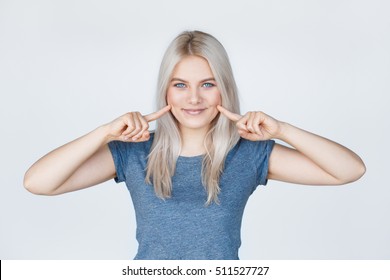 Young Smiling Woman With Fingers Pointing To Her Dimples. Beautiful Blonde Hipster Girl With Blue Eyes On White.