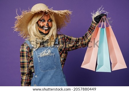 Young smiling woman with Halloween makeup mask in straw hat scarecrow costume hold package bags with purchases after shopping isolated on plain dark purple background studio Celebration party concept