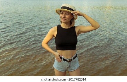 Young smiling woman in a hat and denim shorts on a background of dark water in the river.