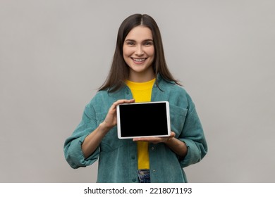 Young smiling woman holding digital tablet in hands, showing blank device screen to camera, expressing happiness, wearing casual style jacket. Indoor studio shot isolated on gray background. - Powered by Shutterstock