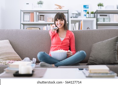 Young Smiling Woman At Home Sitting On The Couch And Watching Tv, She Is Holding A Remote Control And Eating Popcorn