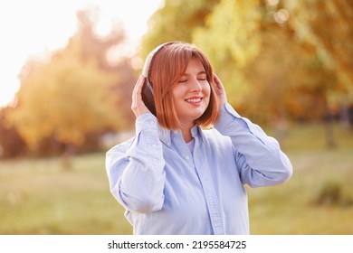 Young smiling woman listening music with headphones at sunset. Caucasian girl listening songs via wireless black headphones. Closeup face of teen and keeps the rhythm of the song.