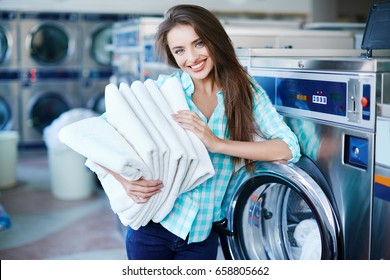 Young smiling woman looking at camera and holding a heap of white clean linen, standing near washing machine. Feeling softness of linen and freshness of washing