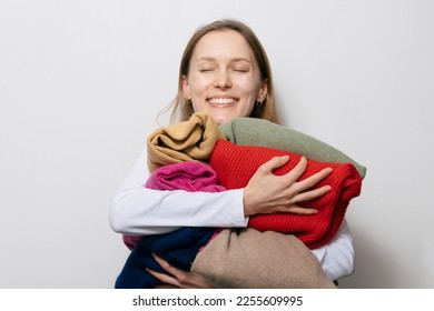 Young smiling woman with loose hair holding and hugging warm winter sweaters of different colors on white isolated background. Concept of changing winter wardrobe and favorite clothes
