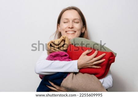 Young smiling woman with loose hair holding and hugging warm winter sweaters of different colors on white isolated background. Concept of changing winter wardrobe and favorite clothes