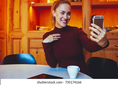 Young smiling woman making photo with smart phone digital camera while sitting in contemporary restaurant interior, happy female is photographing herself on cell telephone during rest in coffee shop