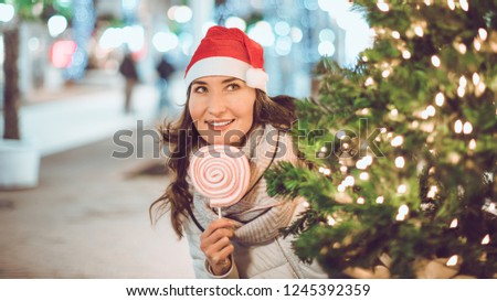 Young smiling woman with marshmallow roller pop in her hand looks out of Christmas tree wearing Santa red hat - winter holidays celebration concept.