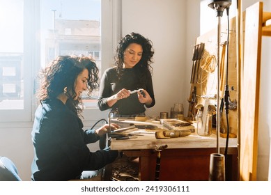 Young smiling woman measuring size of ring with calipers while her twin sister is sitting at wooden workbench and sawing metal in professional jewelry workshop - Powered by Shutterstock