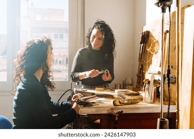 Young smiling woman measuring size of ring with calipers while talking to colleague sitting at wooden workbench in professional jewelry workshop - Powered by Shutterstock