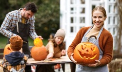 Young Smiling Woman Mother In Apron With Jack O Lantern Smiling At Camera While Carving Pumpkins With Husband And Two Kids In Backyard, Family Preparing Decorations For Halloween Outdoor With Children