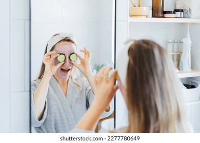 A young smiling woman with pink clay facial mask holds cucumber slices making a refreshing eye mask in bathroom. Natural cosmetic procedures for skin care at home. Beauty self-care. Selective focus - Powered by Shutterstock