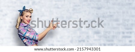 Young smiling woman pointing finger at something. Excited pin up girl, showing advertising some product. Retro fashion vintage concept. White brick wall background. Wide image with ad text copy space.