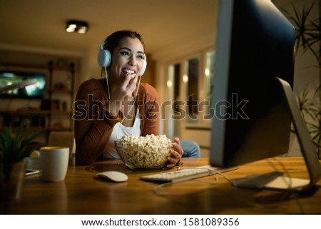 Young smiling woman relaxing in the evening at home while eating popcorn and watching something on a computer. 