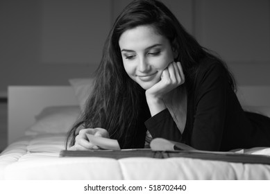 Young Smiling Woman Relaxing On The Bed At Home, She Is Watching Old Pictures On A Photo Album And Lying Down